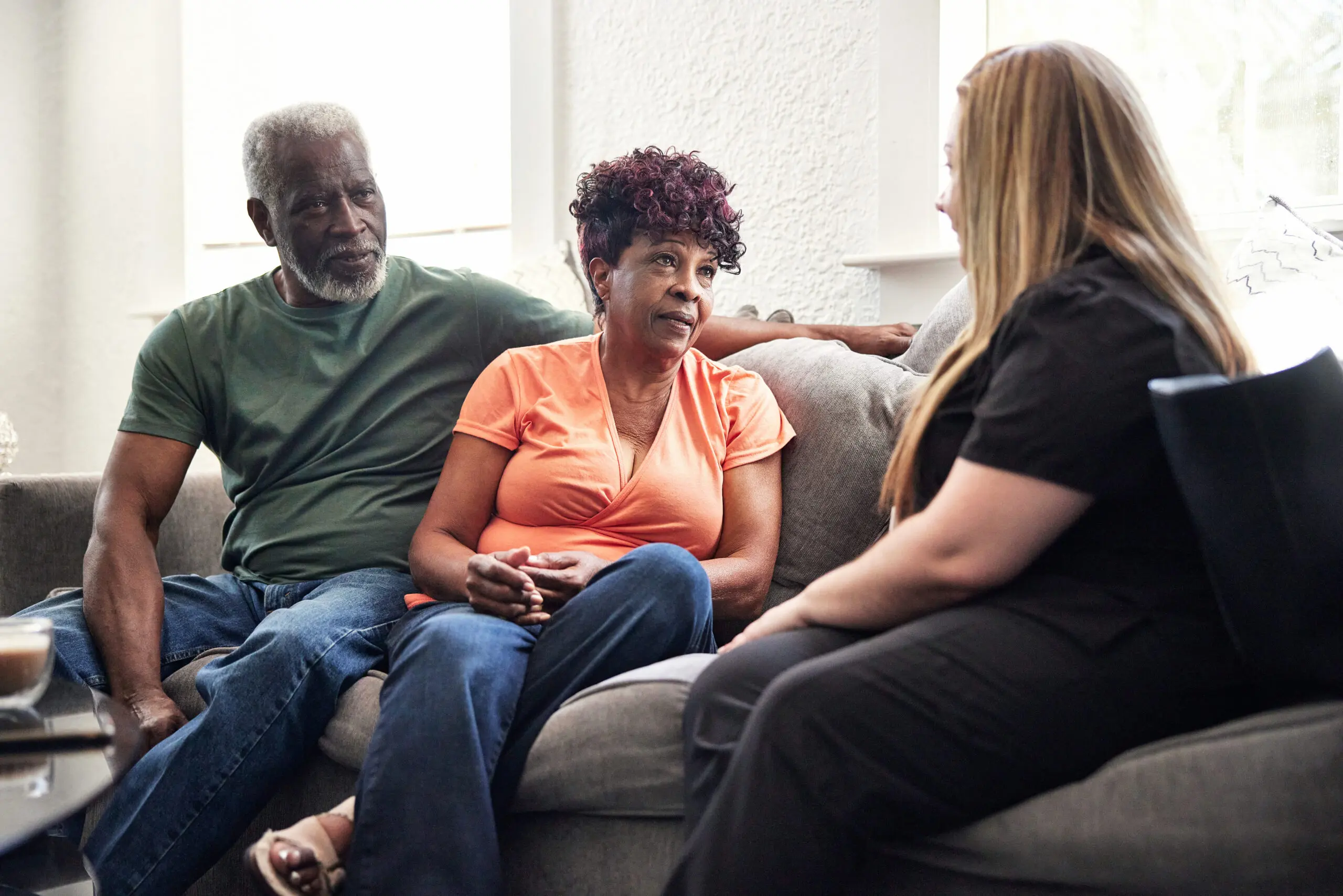 Mid adult healthcare worker in uniform sitting on sofa with Black couple in casual attire and talking about services provided during house calls.